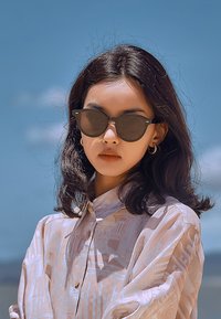 Occhiali da sole di forma rotonda e lenti scure, indossati da una persona in camicia con bottoni a fantasia rosa chiaro contro un cielo blu.