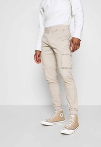 Beige cargo nadrágok elasztikus mandzsettával, sok zsebel, és texturált anyag, bézs magasszárú cipővel párosítva.