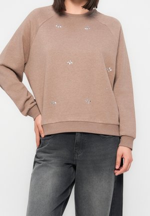 Kvinde iført en beige sweatshirt med små sølvdekorationer og løse mørkegrå jeans, stående med den ene hånd i lommen.