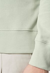 Sudadera verde menta con puños y dobladillo acanalados, fabricada con una tela suave de textura lisa. El puño presenta un detallado pespunte.