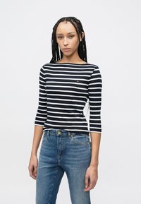 Μπλε ναυτικό και άσπρο ριγέ μακρυμάνικο τοπ με λαιμόκοψη καραβομαστόρικα και λεπτή εφαρμογή, συνδυασμένο με μπλε τζιν παντελόνι με χρυσό κουμπί στο κλείσιμο.