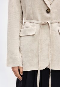 Μπεζ λινό σακάκι με μπροστινές τσέπες με καπάκι, κλείσιμο με ένα κουμπί και ζώνη από το ίδιο ύφασμα, φορεμένο πάνω από μια μαύρη πλισέ φούστα.