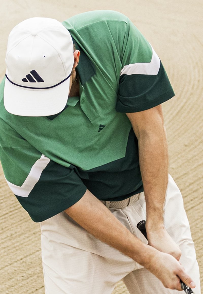 Golfari pelaa vihreässä ja valkoisessa paidassa sekä valkoisessa lippalakkissa, joka heiluttaa mailaa hiekkaisella kentällä.