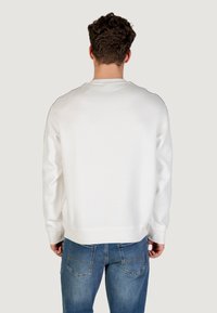 Hombre con cabello corto y rizado, vistiendo un sudadera blanca lisa y jeans azules, de pie de espaldas contra un fondo claro.
