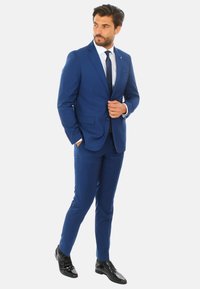 Completo blu su misura con pantaloni slim-fit, revers a dente di cane e due tasche frontali. Abbinato a una camicia bianca e una cravatta scura; scarpe nere.