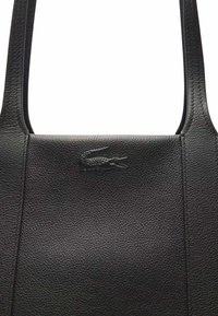 Sac fourre-tout en cuir noir avec une surface texturée, arborant un logo Lacoste en relief. Design simple avec deux anses robustes.