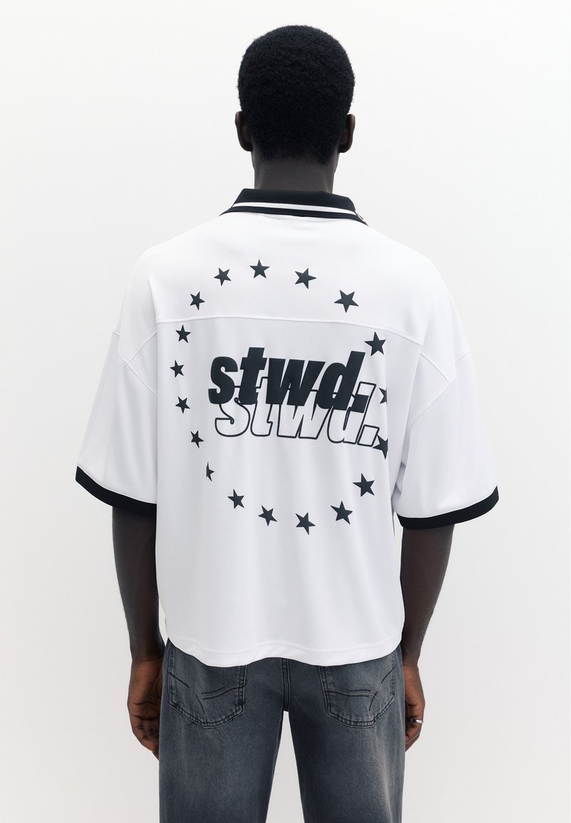 Homme portant une chemise blanche à manches courtes avec col et bordures de manches noirs, étoiles noires et texte « stwd. » dans le dos, associée à un jean foncé.