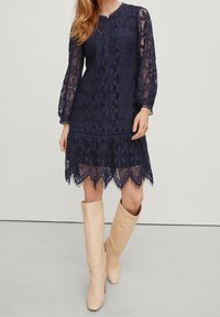 Robe en dentelle marine à manches longues avec un ourlet festonné. Associée à des bottes beiges jusqu'aux genoux. Motifs géométriques détaillés sur l'ensemble.