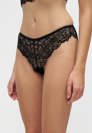 Sous-vêtements en dentelle noire à motifs floraux portés par une personne montrée du milieu du corps jusqu’aux cuisses sur un fond clair uni.