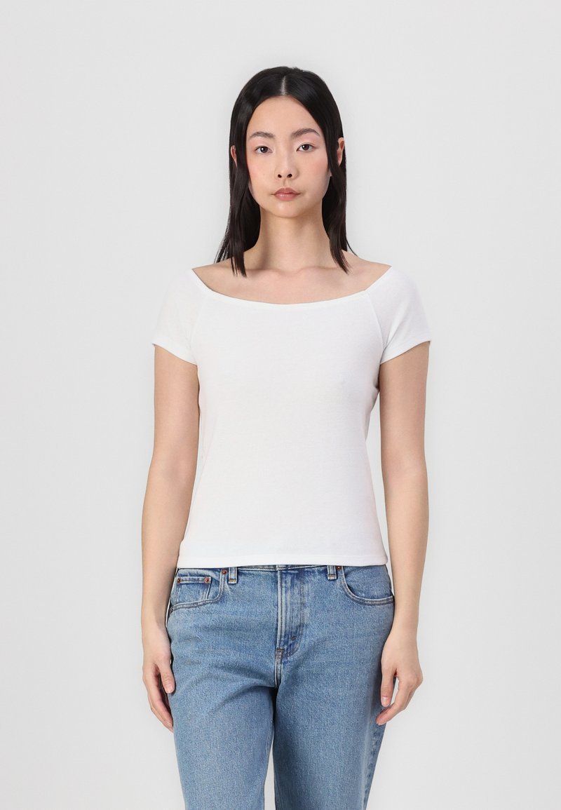 T-shirt branca ajustada com mangas curtas e decote amplo, combinada com jeans de ganga azul claro. Textura suave e design simples.
