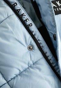 Világoskék steppelt dzseki belseje, fekete cipzárral és "BAKER by TED BAKER" feliratos címkével; látható fémdüces gomb részlet.
