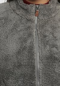 Chaqueta de forro polar gris con cuello alto, cremallera completa y una suave tela texturizada. Incluye un tirador de cuero marrón en la cremallera.