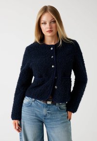 Kobieta z prostymi blond włosami ubrana w granatową, fakturowaną kurtkę z guzikami i jasnoniebieskie dżinsy na jednolitym tle.