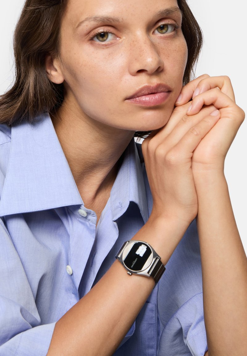 Silberne Metall-Uhr mit schwarzem rundem Ziffernblatt, am Handgelenk getragen, neben einem hellblauen Hemd mit Kragen. Glatte Oberfläche, minimalistisches Design.