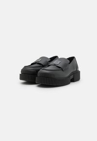 Scarpe slip-on nere realizzate in materiale sintetico liscio, con una suola in gomma prominente caratterizzata da una texture rigata e dettagli con il logo del marchio.