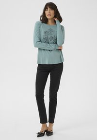 Langarm shirt in teal met een ingewikkeld zwart patroon in het midden, gecombineerd met zwarte slim-fit broek en zwarte flats met strikdetail. Gladde textuur.
