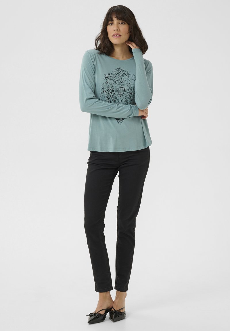 Langarm shirt in teal met een ingewikkeld zwart patroon in het midden, gecombineerd met zwarte slim-fit broek en zwarte flats met strikdetail. Gladde textuur.