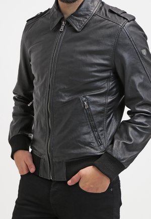 Homme portant une veste en cuir noire avec fermeture éclair à l'avant et poches, poignets et ourlet côtelés, les mains partiellement dans les poches.