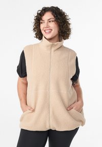 Beige fleece vest met hoge kraag, rits aan de voorkant en zijzakken. Zachte, gestructureerde stof, korte mouwen gelayerd over een zwart shirt.