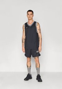 Camisola de basquetebol preta com ribete branco, combinada com calções de rede pretos e sapatilhas escuras. Meias cinzentas até ao joelho. Tatuagens visíveis nos braços.