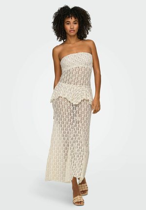 Kobieta o kręconych włosach, ubrana w beżową, koronkową suknię maxi bez ramiączek i beżowe plecione sandały, stojąca na białym tle.