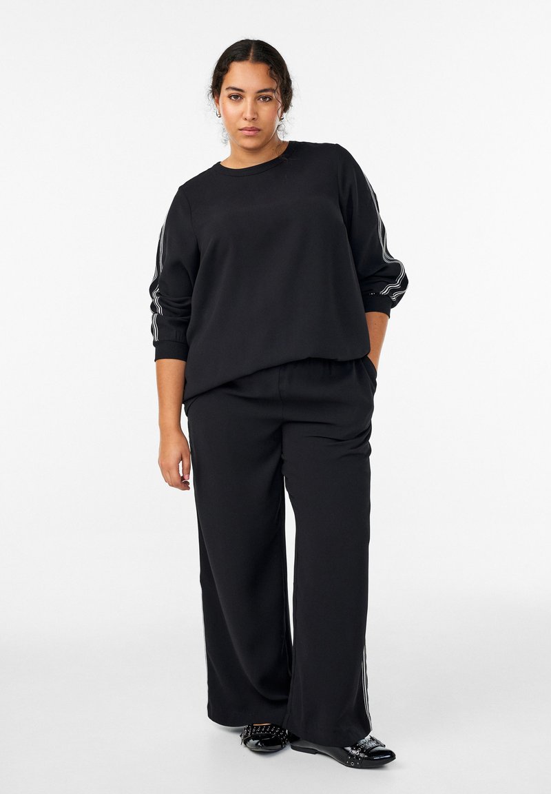 Žena stojaca s jednou rukou v vrecku, oblečená v čiernom tričku s dlhým rukávom a širokých nohaviciach s bočnými pruhmi a čiernymi zdobenými topánkami.