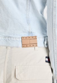 Světle modrá denimová bunda s hnědou koženou nášivkou s nápisem "TOMMY JEANS" našitou na zadní straně; doplněna světle béžovými kalhotami s boční kapsou.