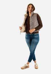 Chaleco beige con cuello y panel de lunares de leopardo, llevado sobre un suéter marrón texturizado y pantalones vaqueros azules. Zapatillas beige y marrones con cordones.