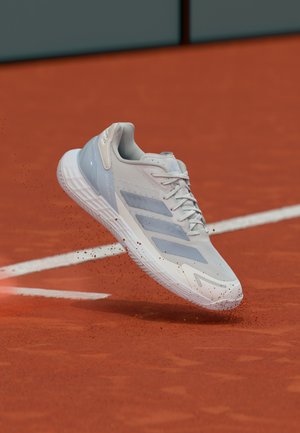 Zapatilla deportiva blanca y azul claro con parte superior texturizada, tres rayas diagonales y suela flexible, mostrada en el aire sobre una pista de arcilla roja.