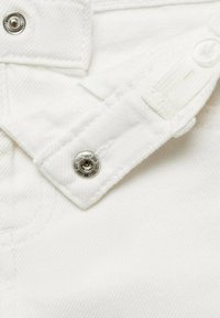 Detail blízko bílého denimu, který ukazuje knoflík a nastavitelné popruhy, s texturovaným povrchem a kovovými akcenty knoflíků.