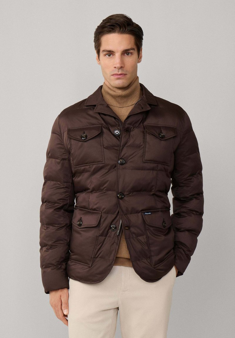 Chaqueta acolchada marrón con diseño aislante, que cuenta con dos bolsillos en el pecho, dos bolsillos frontales y cierres de botones. Se lleva sobre un suéter de cuello alto beige.