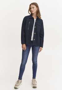 Marineblå denimjakke med krage, frontlomme med glidelås og gullknapper. Brukt over en hvit t-skjorte, kombinert med blå skinny jeans og joggesko.