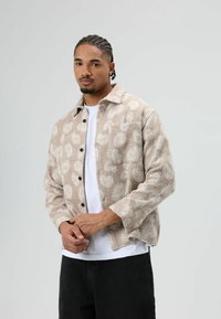 Beige overhemdjas met patroon en knoopsluiting, voorzien van een zak op de linkerborst en zichtbare naden. Gedragen over een wit overhemd.