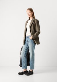 Oliwkowa marynarka na białej teksturowanej bluzce, zestawiona z jasnoniebieskimi prostymi dżinsami i czarnymi masywnymi loafersami. Obecna jest czarna torba.