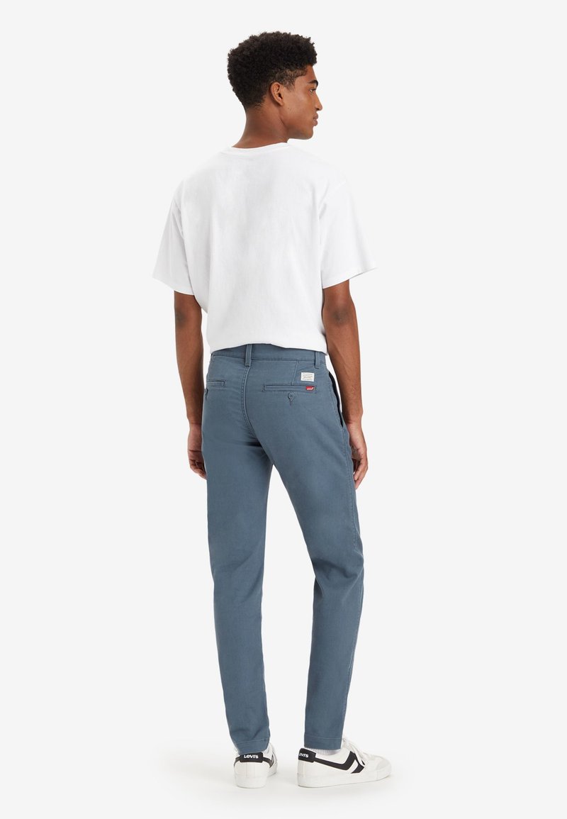Chinos bleus pour hommes avec une coupe slim, fabriqués en coton. Présentent des poches avant et arrière, ainsi qu'une étiquette sur la poche arrière. Associés à un t-shirt blanc.