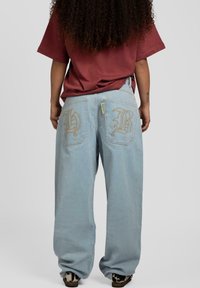Lyseblå baggy jeans med broderte gullbokstaver på baklommene, paret med en maroon oversized t-skjorte.