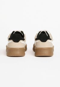Beige nahkasneakerit mustilla yksityiskohdilla, karkeapintainen ruskea kumipohja, pyöreä muoto ja takana vetolenkit helppoa pukemista varten.