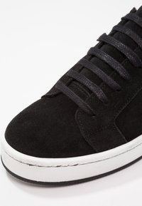 Gros plan d'une basket en daim noir avec des lacets noirs et une semelle en caoutchouc blanche sur fond blanc.