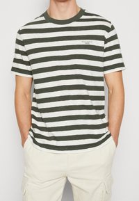 Hombre que lleva una camiseta de rayas horizontales verde oscuro y blanca de manga corta, y pantalones cargo de color beige claro, con las manos en los bolsillos.