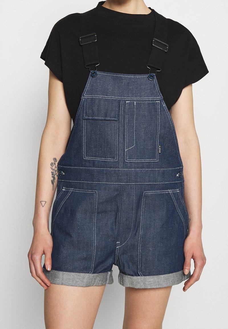 Personne portant une salopette courte en denim bleu foncé avec des poches avant et des bretelles noires réglables sur un t-shirt noir à manches courtes.