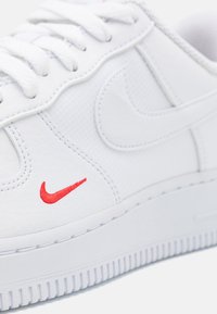 Sneaker bianca realizzata in materiale sintetico testurizzato. Caratterizzata da un logo ricamato rosso, lacci bianchi piatti e una suola spessa con scanalature per il supporto.