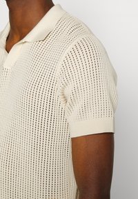 Dzianinowa koszulka polo w kolorze kremowym, z otwartym wzorem ażurowym, krótkimi rękawami oraz standardowym kołnierzykiem z prążkowanymi detalami.