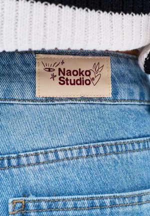Dżinsy z denimu z beżową metką z napisem "Naoko Studio", na której znajduje się oko, gwiazdy i serce z liśćmi, przyszyte na tylnej części pasa.