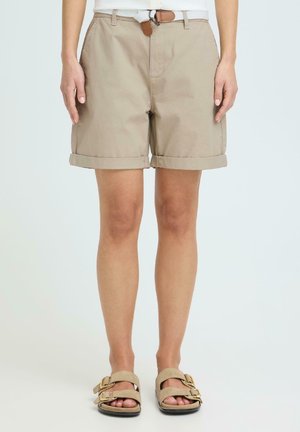 Person iført beige shorts med oppbrettede kanter, brun belte og beige sandaler med spenne, stående mot en ensfarget lys bakgrunn.
