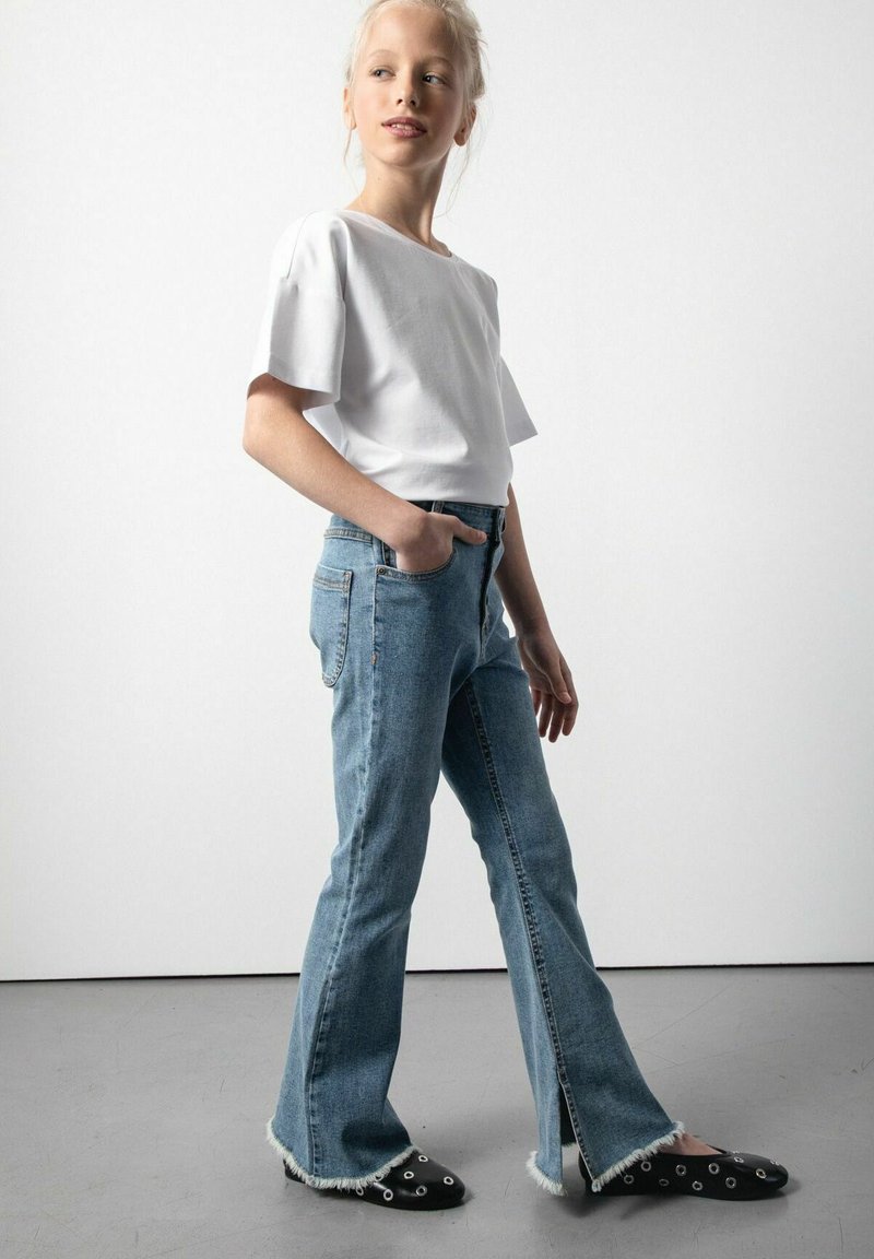 Biała koszulka z krótkim rękawem, jasnoniebieskie dżinsy z szerokimi nogawkami i rozcięciami z przodu oraz czarne buty wsuwane z ozdobnymi dziurkami. Neutralne tło.