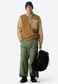 Fleecová mikina v hnědé a béžové barvě s předním zipem a zelenou kapsou; doplněná zelenými cargo kalhotami, černým batohem, modrou čepicí a černými botami.