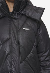 Μαύρο μπουφάν με γυαλιστερή εμφάνιση, ραβδωτό σχέδιο, ψηλό γιακά και λογότυπο στο στήθος. Κλείσιμο με φερμουάρ και υφάσμα με υφή.