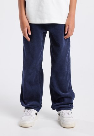Kind dat losse donkerblauwe corduroy broeken en witte sneakers draagt, staande tegen een effen achtergrond.