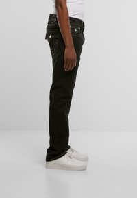 Jeans de denim negro con ajuste slim, con costuras en contraste, cintura de media altura y diseño de cinco bolsillos. Combinados con zapatillas blancas.
