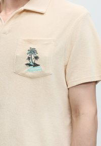 Polo beige à manches courtes avec une poche devant brodée de petits palmiers et un mot bleu clair en dessous.